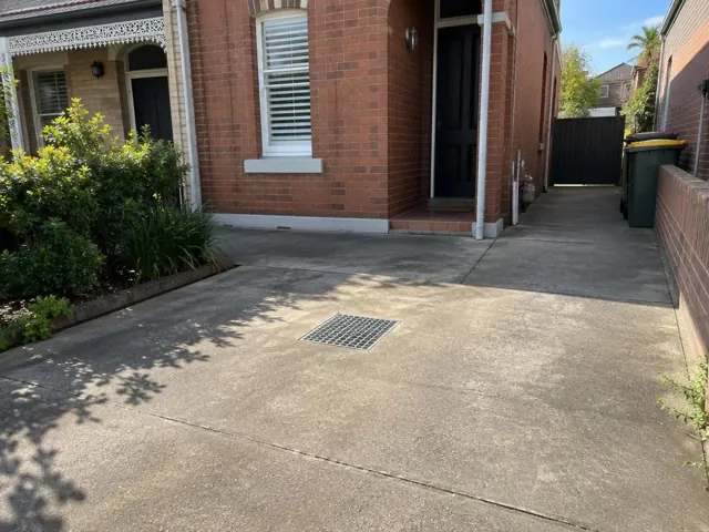 Dry paved driveway with a square drain grate beside a brick house in Clovelly
